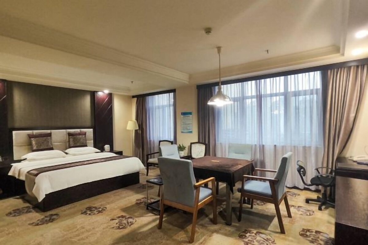 A hotel room with a bed, a dining table with chairs, a desk, a lamp, and a large window with curtains.