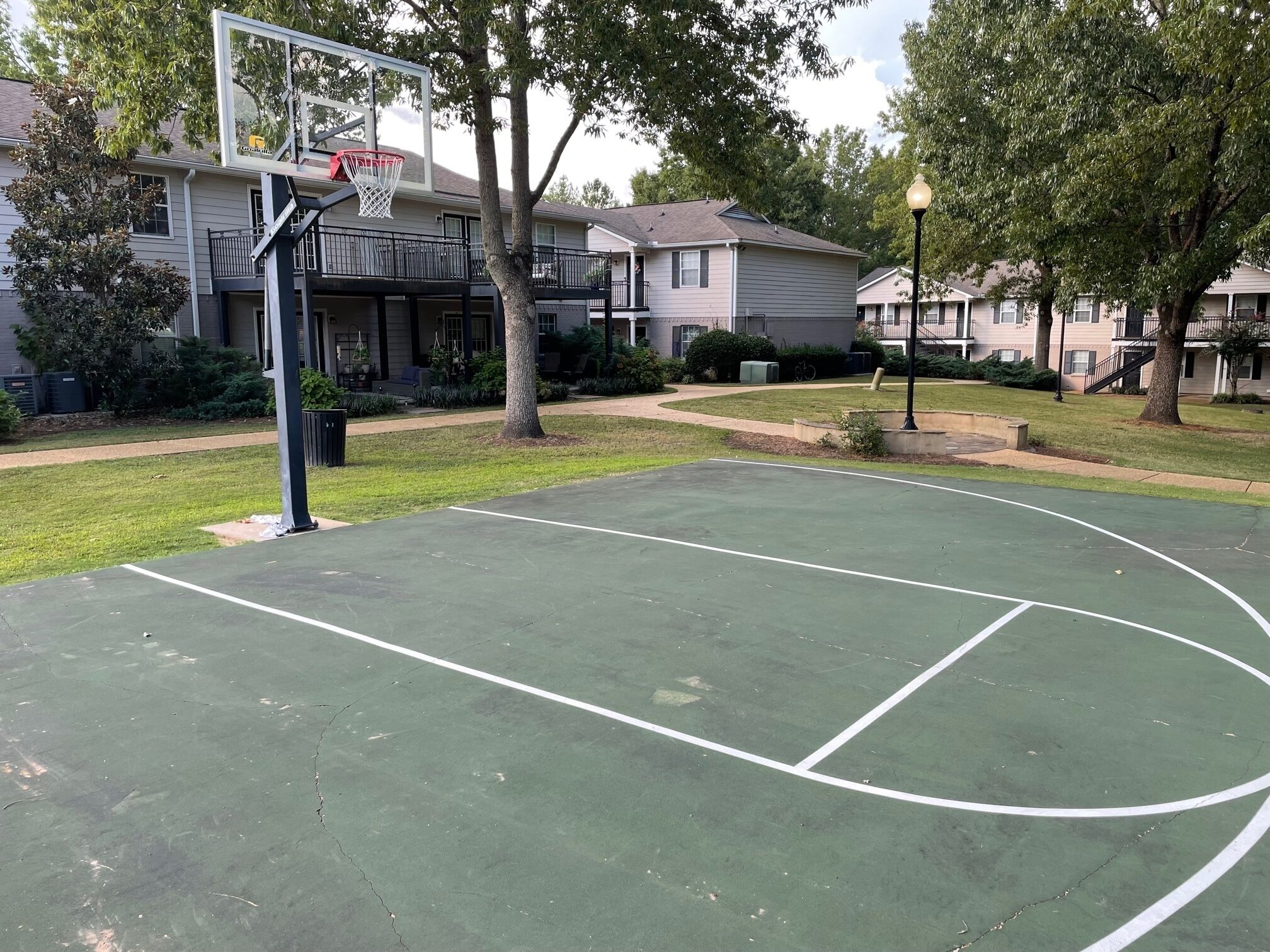 Uma quadra de basquete ao ar livre com uma cesta, cercada por árvores e edifícios residenciais.