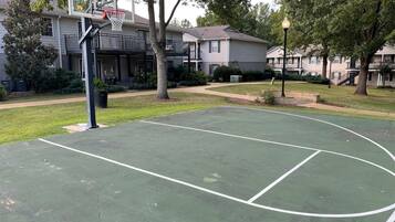 Uma quadra de basquete ao ar livre com uma cesta, cercada por árvores e edifícios residenciais.