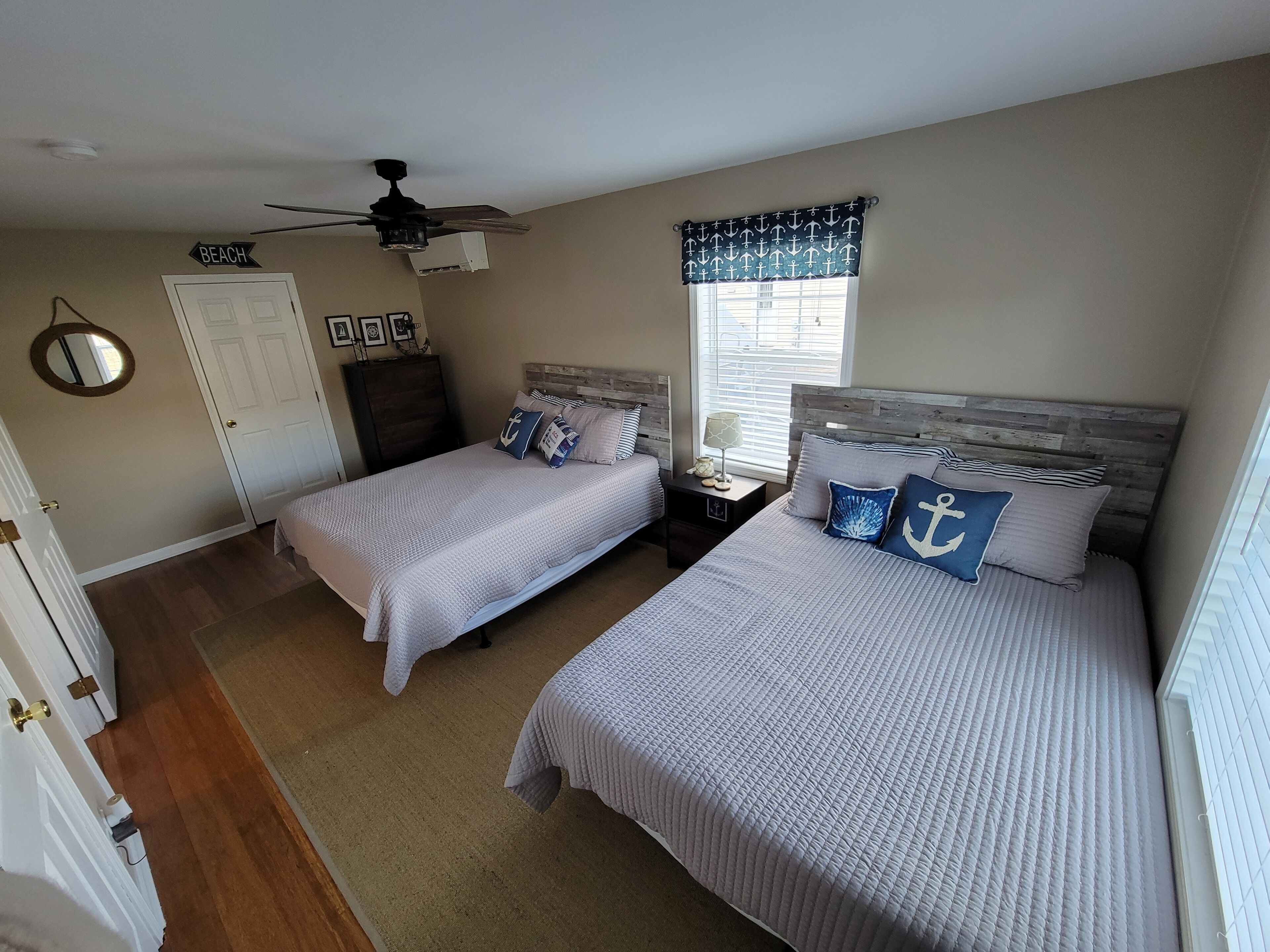 Une chambre avec deux lits, un ventilateur de plafond, une fenêtre avec des rideaux à motifs d’ancres et une porte portant l’inscription « PLAGE ».