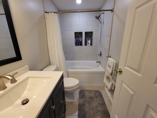 A bathroom with a white sink, toilet, and bathtub. There is a shower curtain, a towel rack, and a small window with bottles on the wall. - PRICE DROP - HIGHLY RATED! Cozy, single family home close to beach and golf (Ocean View)