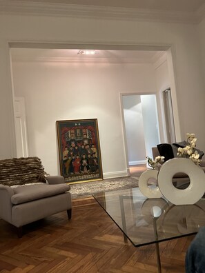 Un salon comprenant un canapé, une table basse en verre, un grand tableau encadré et une porte donnant sur une autre pièce.