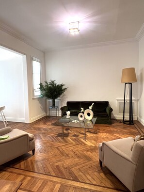 Uma sala de estar com piso de madeira em padrão herringbone, um sofá verde, duas poltronas, uma mesa de centro de vidro com objetos decorativos, um abajur alto e uma planta em vaso.