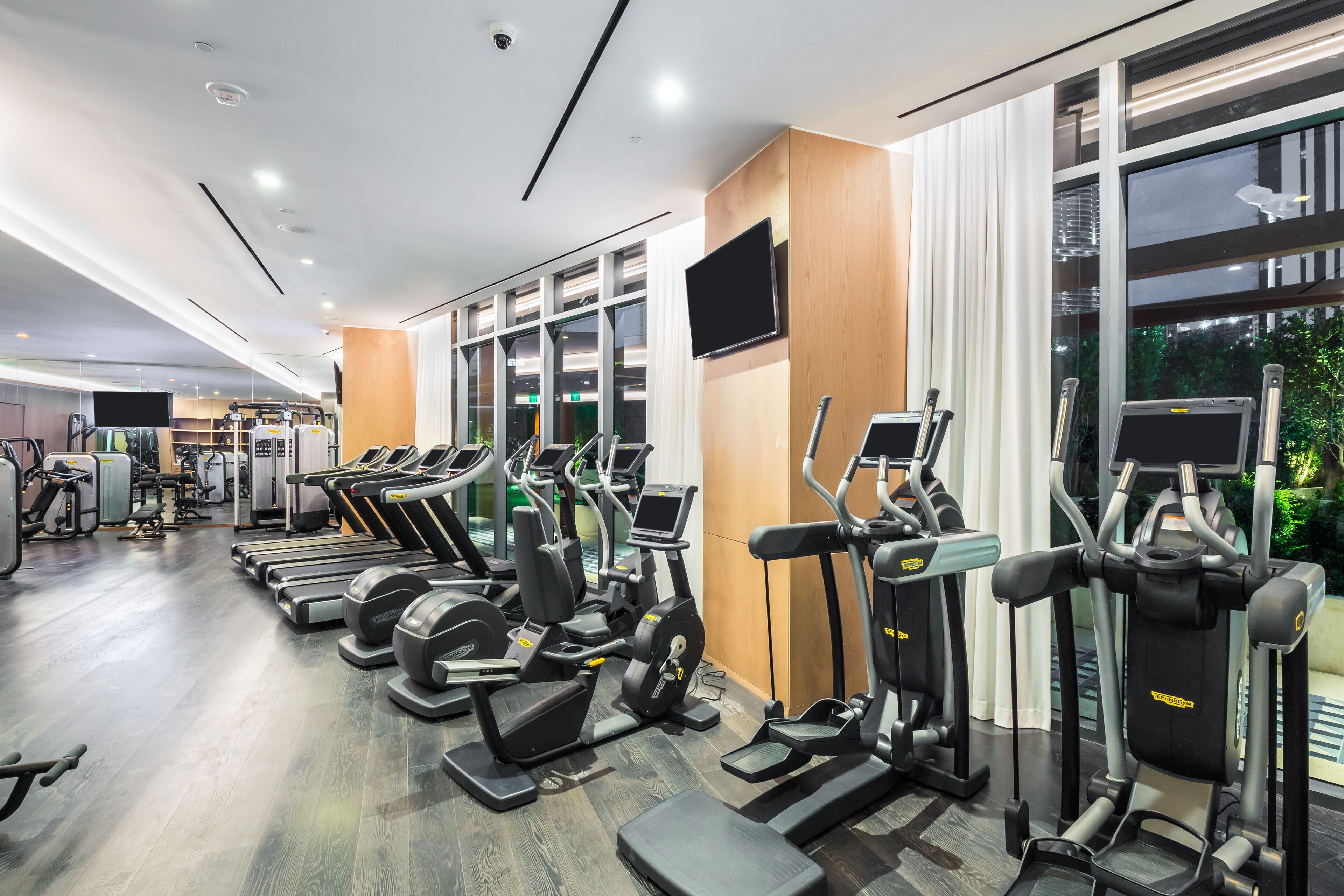 Ein modernes Fitnessstudio mit verschiedenen Cardiogeräten, einem Fernseher und großen Fenstern mit Blick ins Freie.