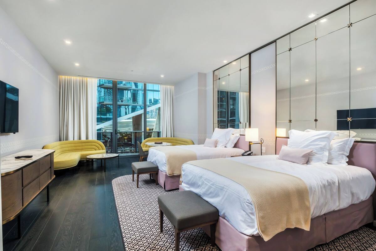A modern hotel room with two beds, a sofa, a desk, and a large window offering a city view.