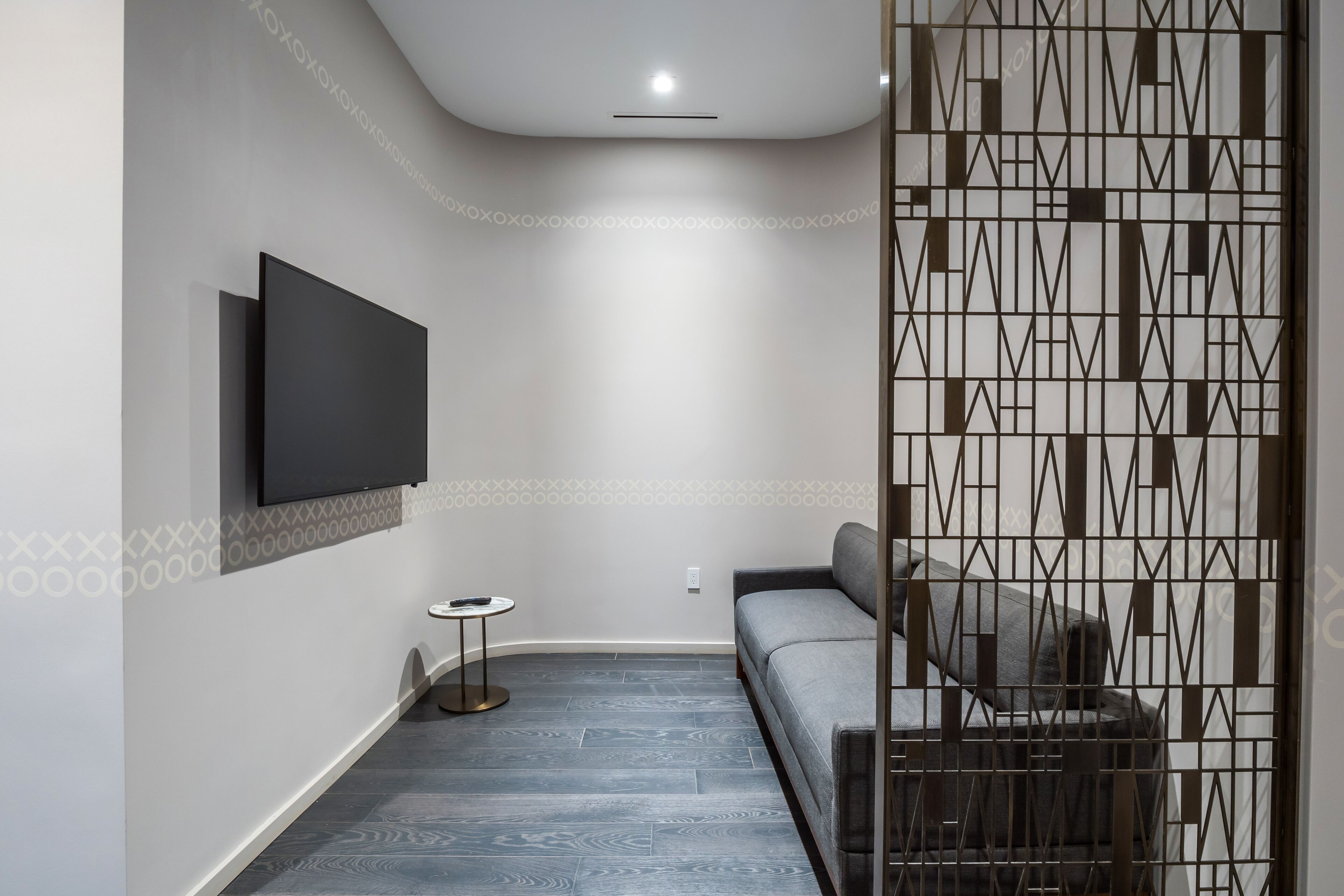 A modern living room with a flat-screen TV mounted on the wall, a grey sofa, a small round side table, and a patterned partition.