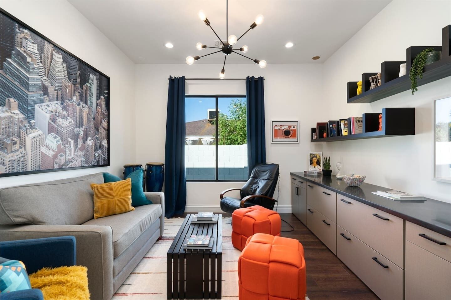 A modern living room with a cityscape painting, a sofa with colorful cushions, a black coffee table, an orange ottoman, a black leather chair, and a kitchen area with a countertop and shelves.