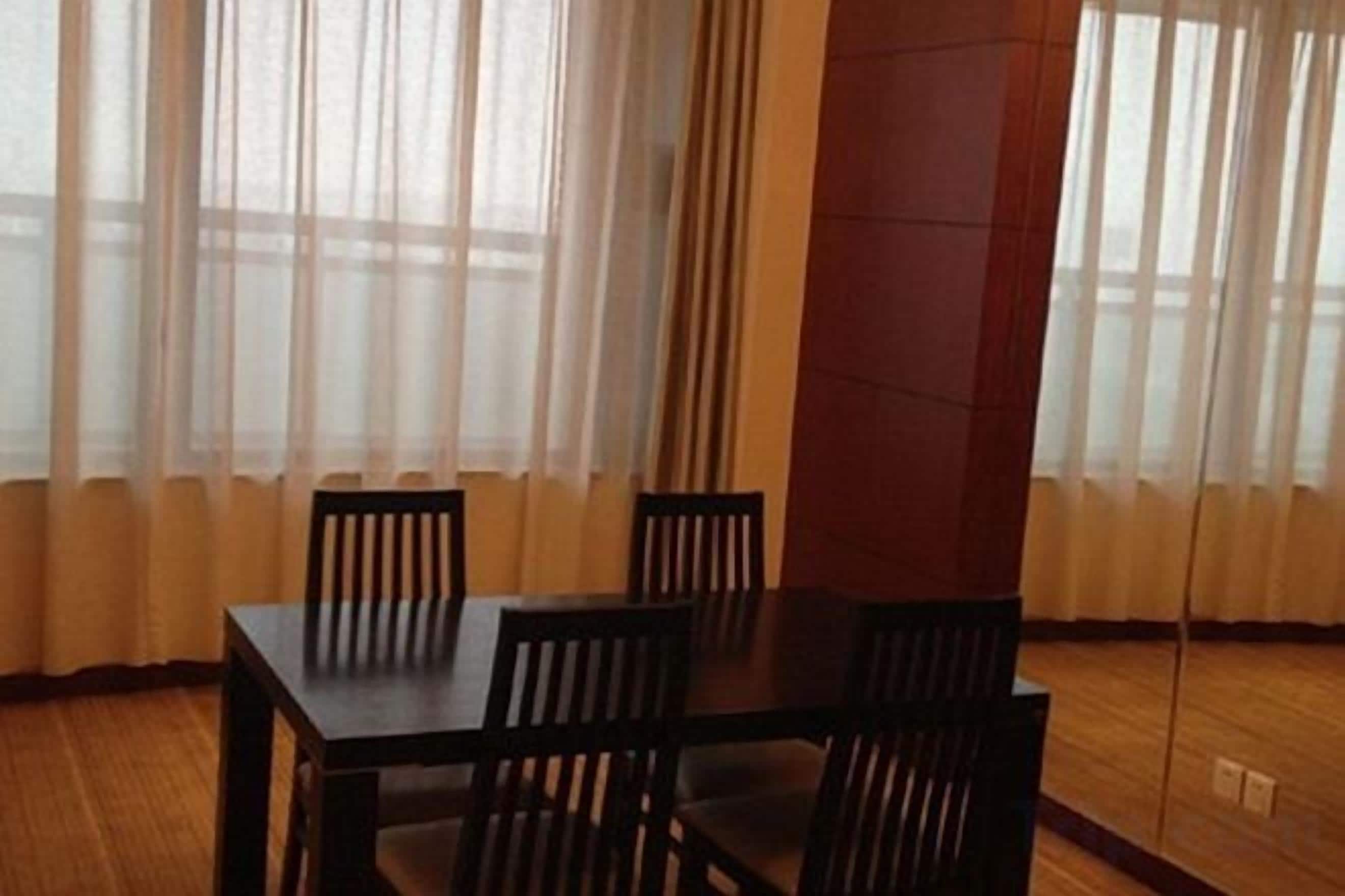 A dining area with a table and chairs, large windows with sheer curtains, and a wooden floor.