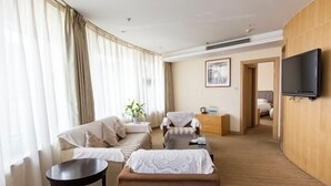 A modern hotel room with a sofa, armchair, and a small table. There is a television mounted on the wall and a window with curtains. - University Hotel (Binzhou)