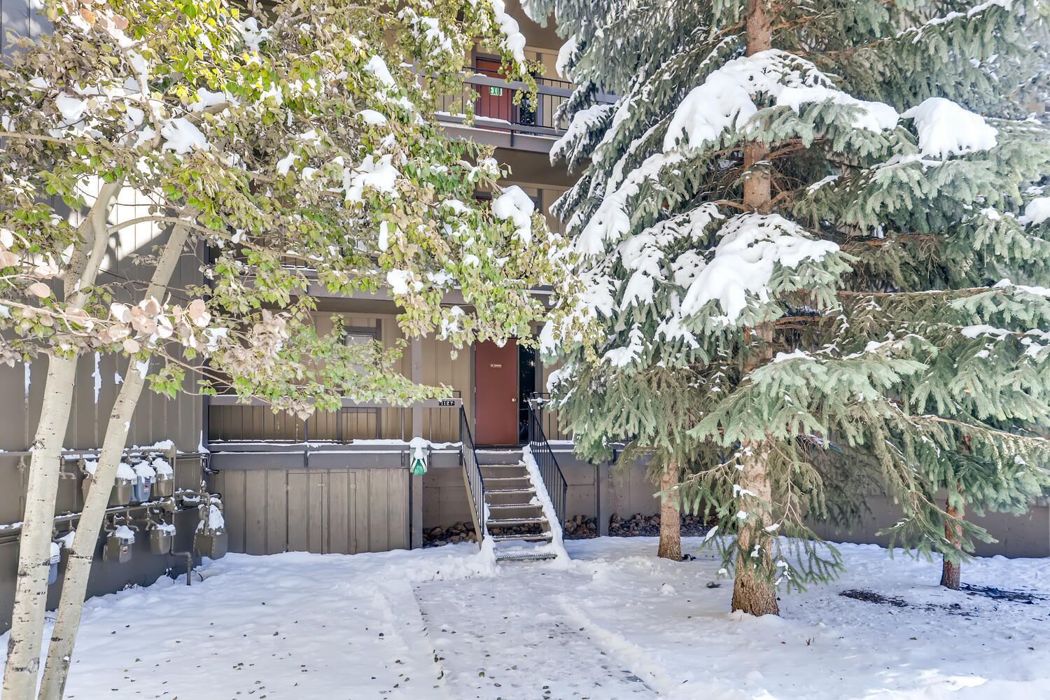 A snow-covered outdoor area with a staircase leading to a building entrance, surrounded by trees and a fence.