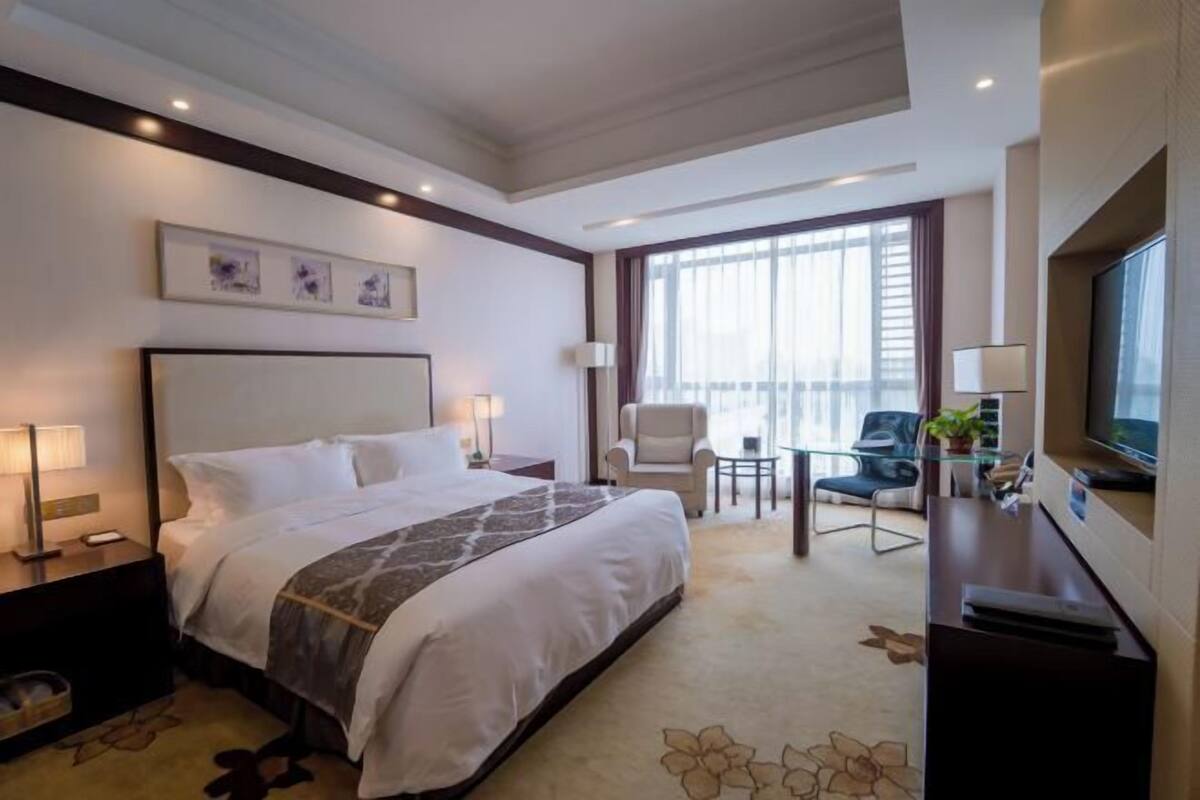 Habitación de hotel con una cama grande, un escritorio con televisor, una silla y un balcón con vistas.