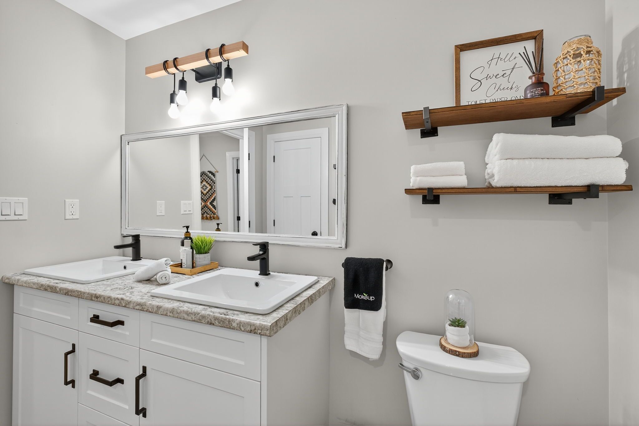 Une salle de bain moderne avec un meuble-lavabo à deux vasques, un grand miroir, des toilettes et une étagère contenant des articles de toilette.