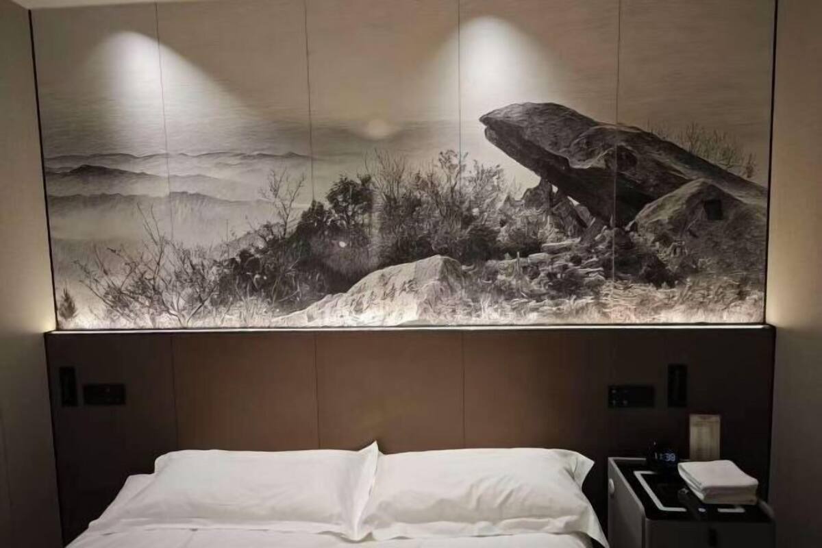 A hotel room with a large wall mural depicting a mountainous landscape, a bed with white linens, and a bedside table with a lamp and a folded towel.