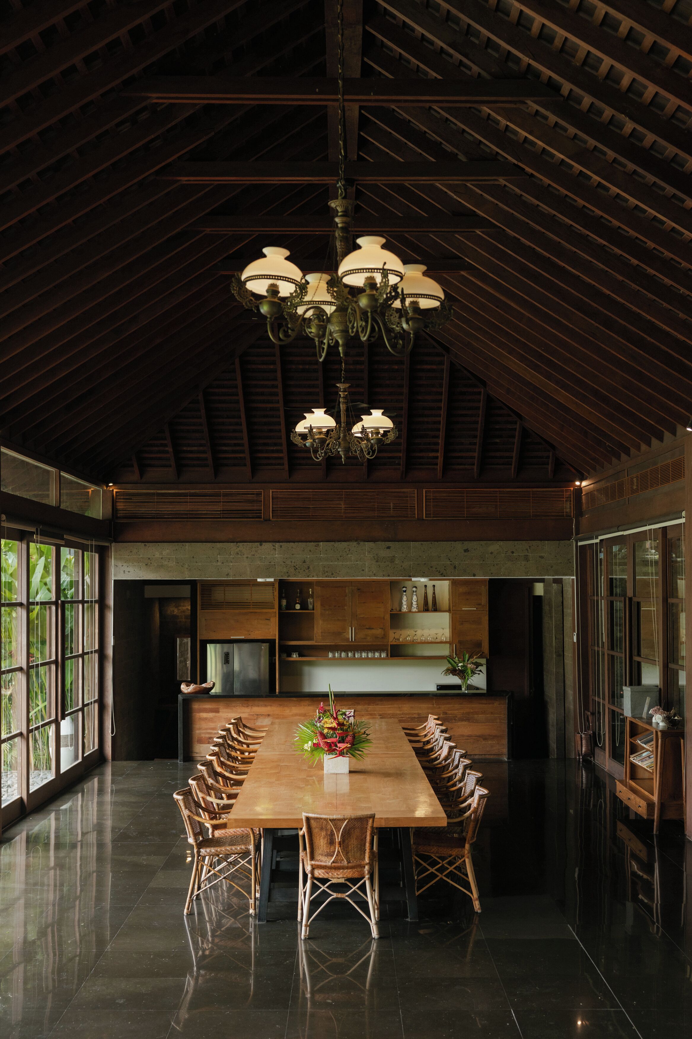 A spacious dining area with a long wooden table and wicker chairs, a chandelier, and a kitchen area with wooden cabinets and a refrigerator.