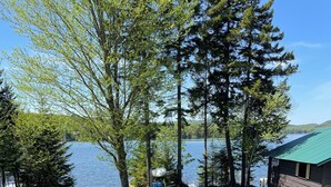 A wooden deck with a table and chairs, overlooking a lake with trees and a cabin. - Lakefront Cabin on Back Lake w trail access, and private dock. (Pittsburg)