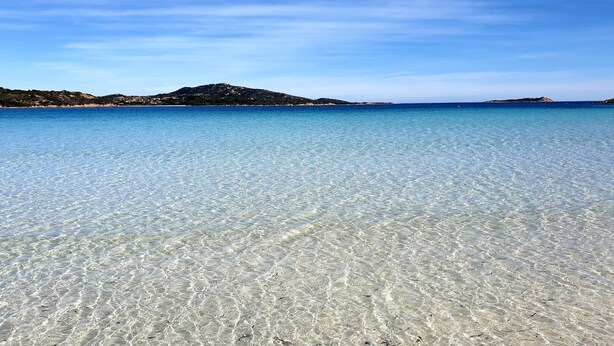 Ein flach abfallender Strand mit seichtem Wasser und Hügeln im Hintergrund.