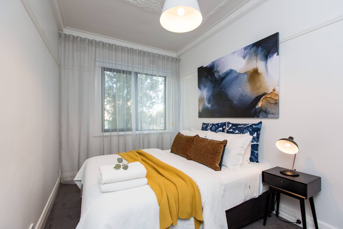 Une chambre à coucher avec un lit, une table de chevet, une lampe, une fenêtre avec des rideaux et un tableau au mur.