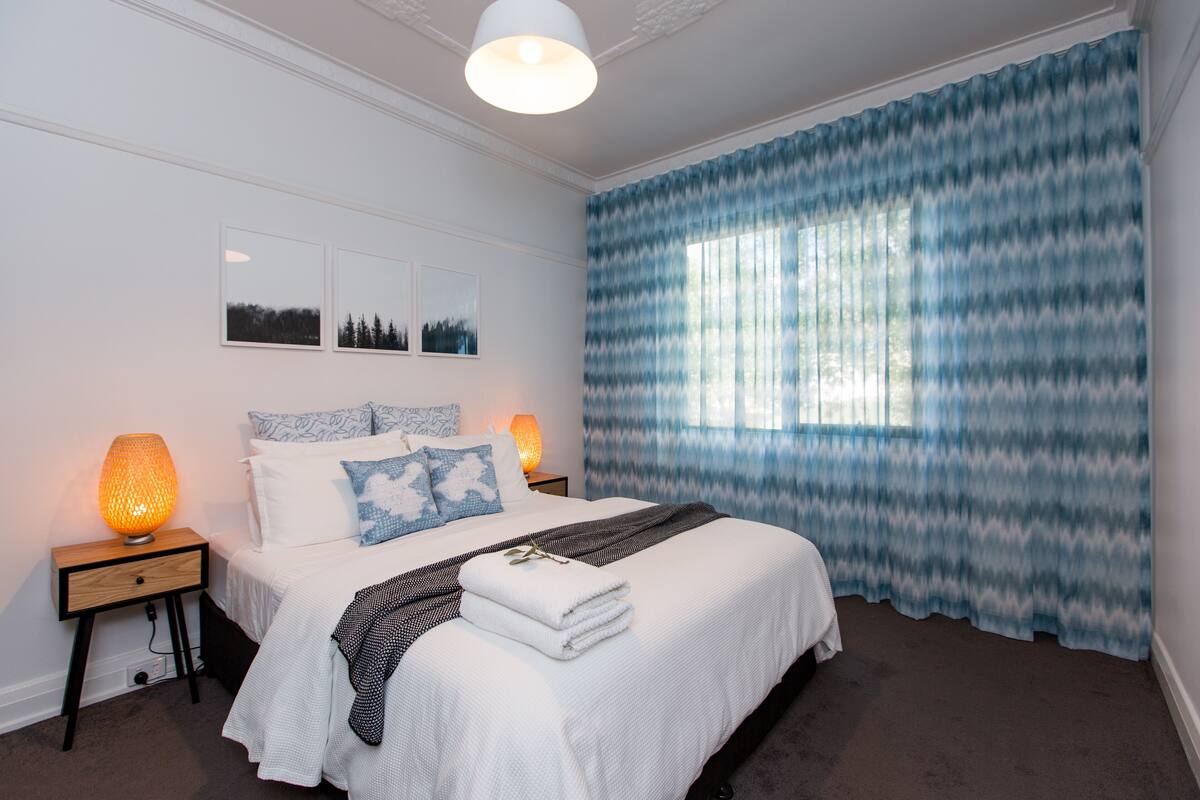 Une chambre à coucher comprenant un lit, deux tables de chevet, une lampe, une fenêtre avec des rideaux et des œuvres d’art murales.