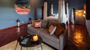 A modern bedroom with a four-poster bed, a sofa, a coffee table with a candle, and a mural on the wall. - Balcones 5BR Villa | Weddings | Stunning Views (San Miguel de Allende)