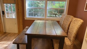 Een houten eettafel met een bank, een rieten stoel en een raam met uitzicht op groen.