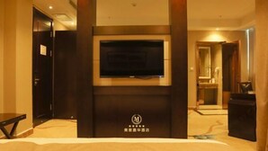A hotel room with a large flat-screen TV mounted on a dark wooden panel, a bed with a beige headboard, and a small table. - Meijia Jiahua Hotel (Shangzhi)