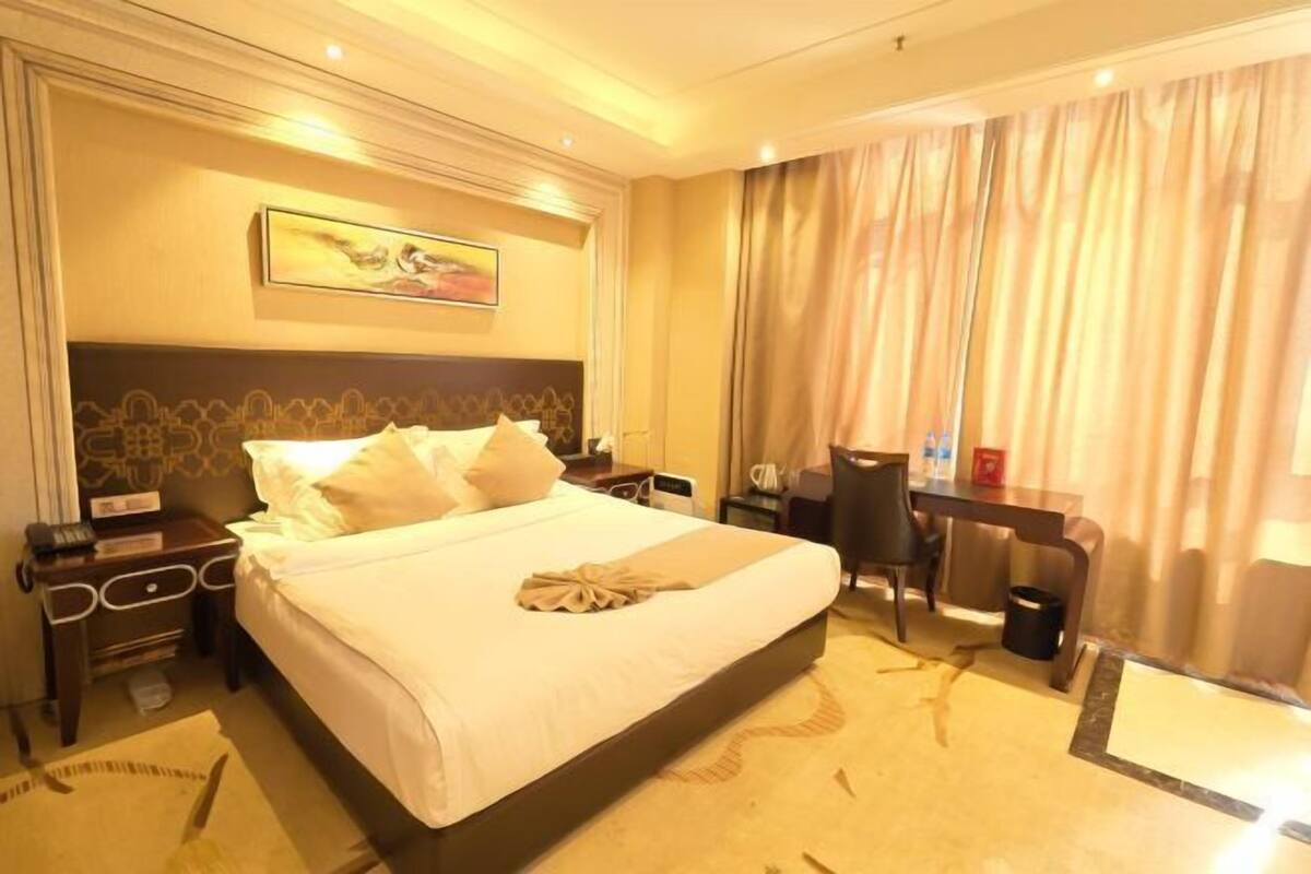 A hotel room with a large bed, a desk, and a chair. There is a painting on the wall and curtains on the window.