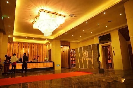 A hotel lobby with a reception desk, a chandelier, a red carpet, and a wall with a large abstract design.. Meijia Jiahua Hotel