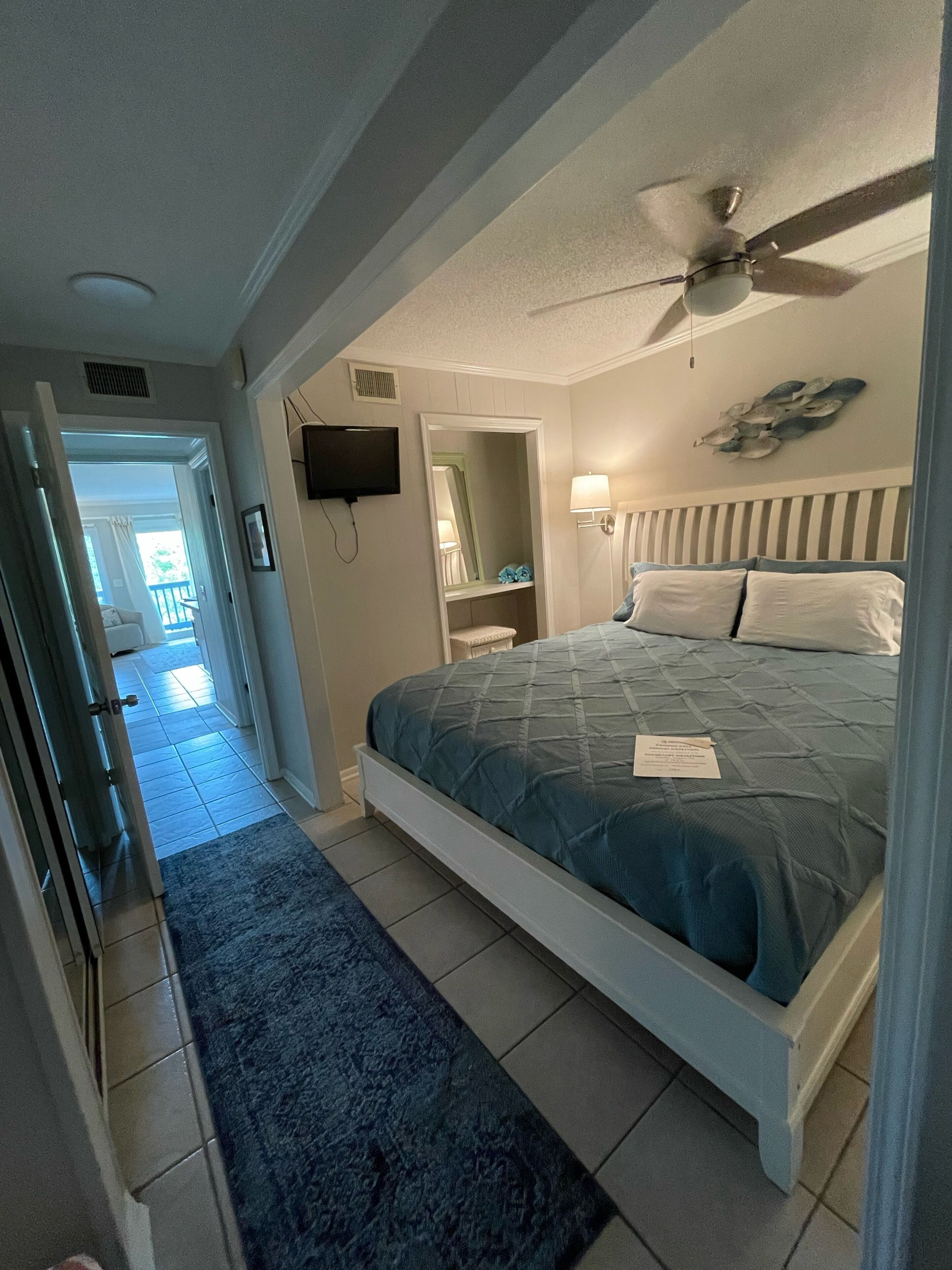 Een slaapkamer met een bed, een plafondventilator, een televisie en een zichtbare badkamer door een geopende deur.