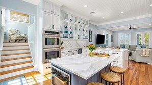 A modern kitchen with white cabinetry, a large island, and a dining area with bar stools. - Stunning Marsh Views, 5 Bedroom 6.5 Bath with pool (Saint Simons Island)