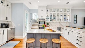 A modern kitchen with white cabinetry, a marble island, and wooden stools. - Stunning Marsh Views, 5 Bedroom 6.5 Bath with pool (Saint Simons Island)