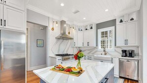 A modern kitchen with white cabinetry, a marble island, stainless steel appliances, and a central island with a floral arrangement. - Stunning Marsh Views, 5 Bedroom 6.5 Bath with pool (Saint Simons Island)