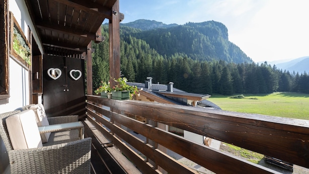 Een houten balkon met rieten meubilair, een zwarte deur met hartvormige spiegels, en uitzicht op een bebost gebergte.