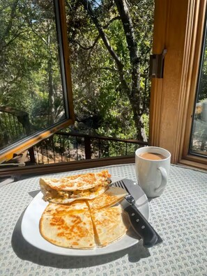 Une table dressée pour le petit-déjeuner, avec une assiette de quesadillas et une tasse de café, placée près d’une fenêtre offrant une vue sur les arbres.