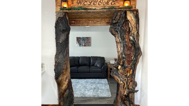 Ein rustikaler Holzbogen mit einem 'WELCOME'-Schild und zwei Laternen, der zu einem Wohnzimmer mit einem schwarzen Sofa und einem gerahmten Bild an der Wand führt.