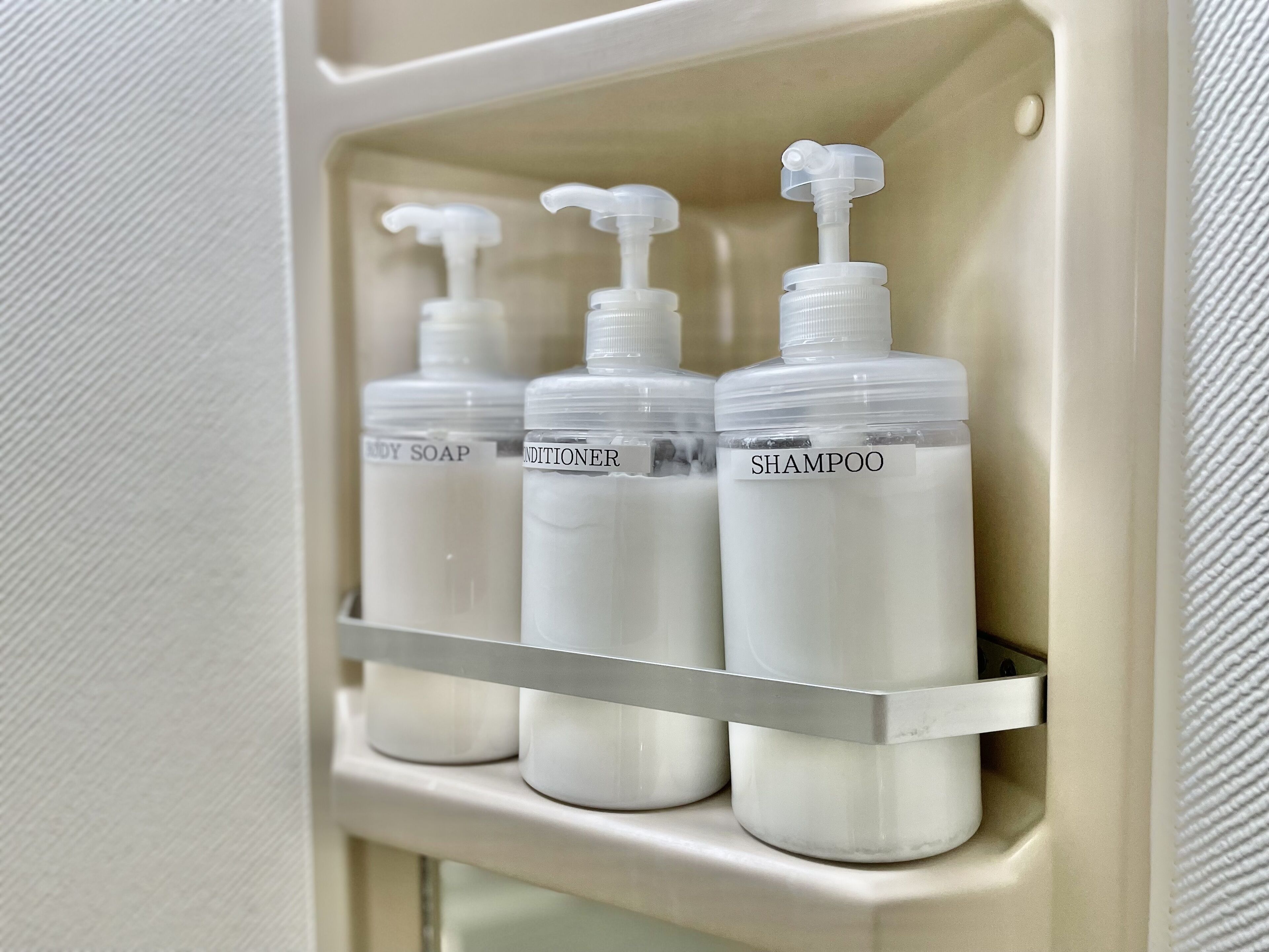 A bathroom shelf with three bottles labeled body soap, conditioner, and shampoo.