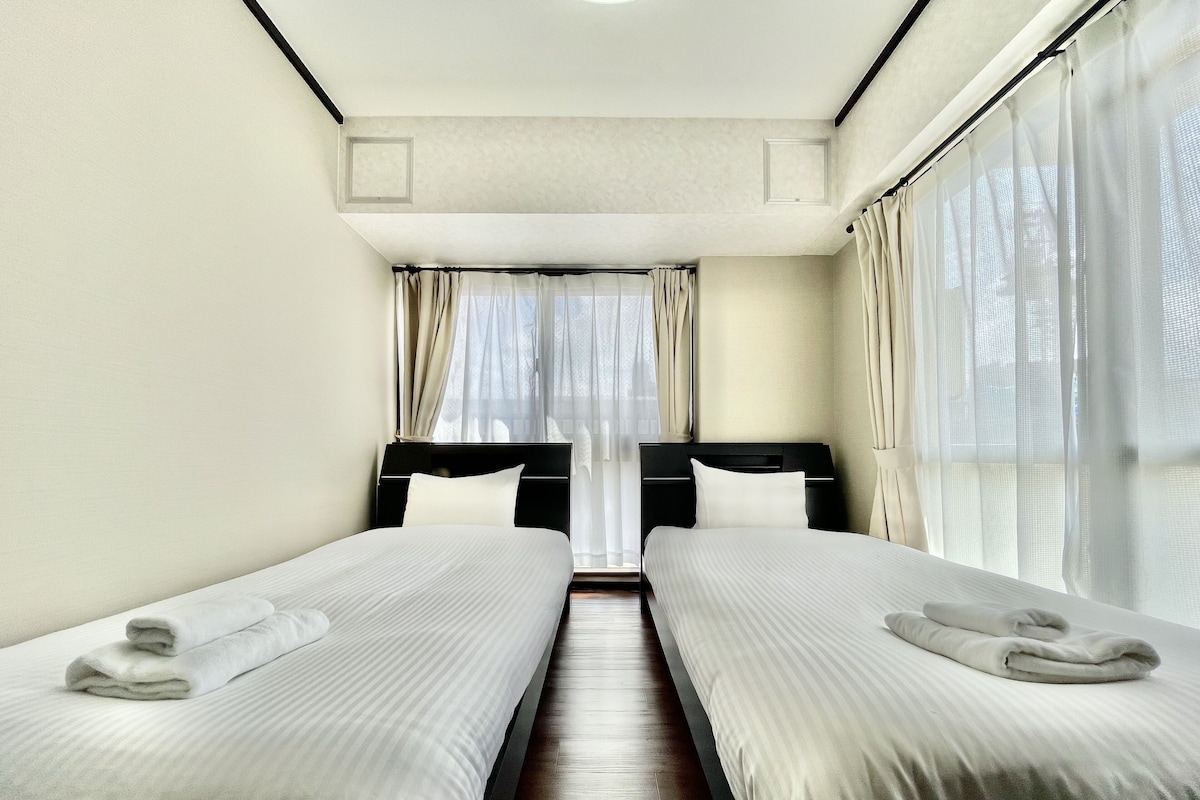 A hotel room with two single beds, a wooden floor, and large windows with sheer curtains.