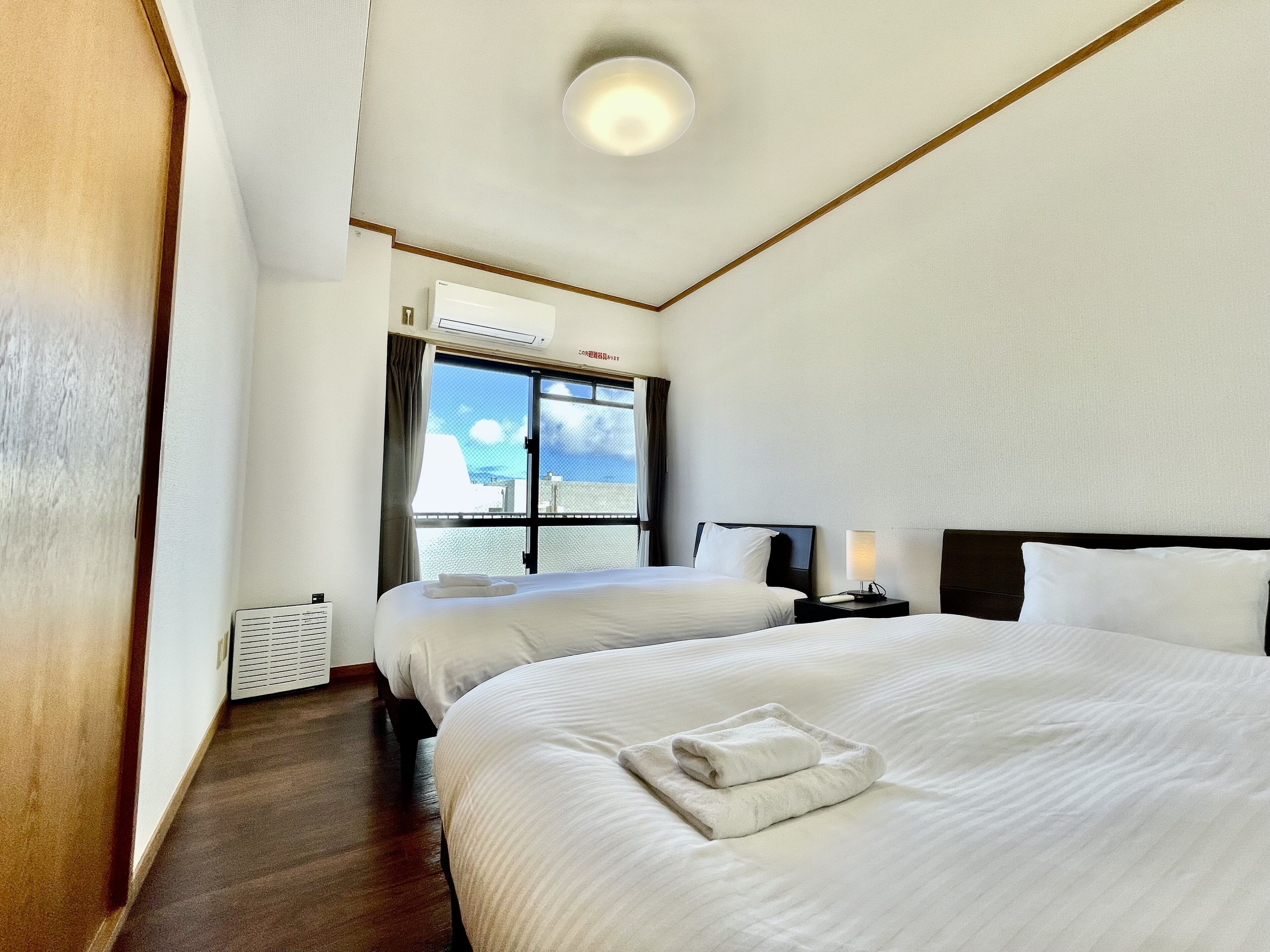 A hotel room with two beds, a window with a view, an air conditioning unit, and a bedside table with a lamp.