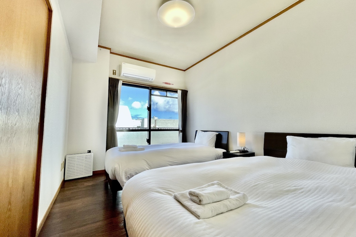 A hotel room with two beds, a window with a view, an air conditioning unit, and a bedside table with a lamp.