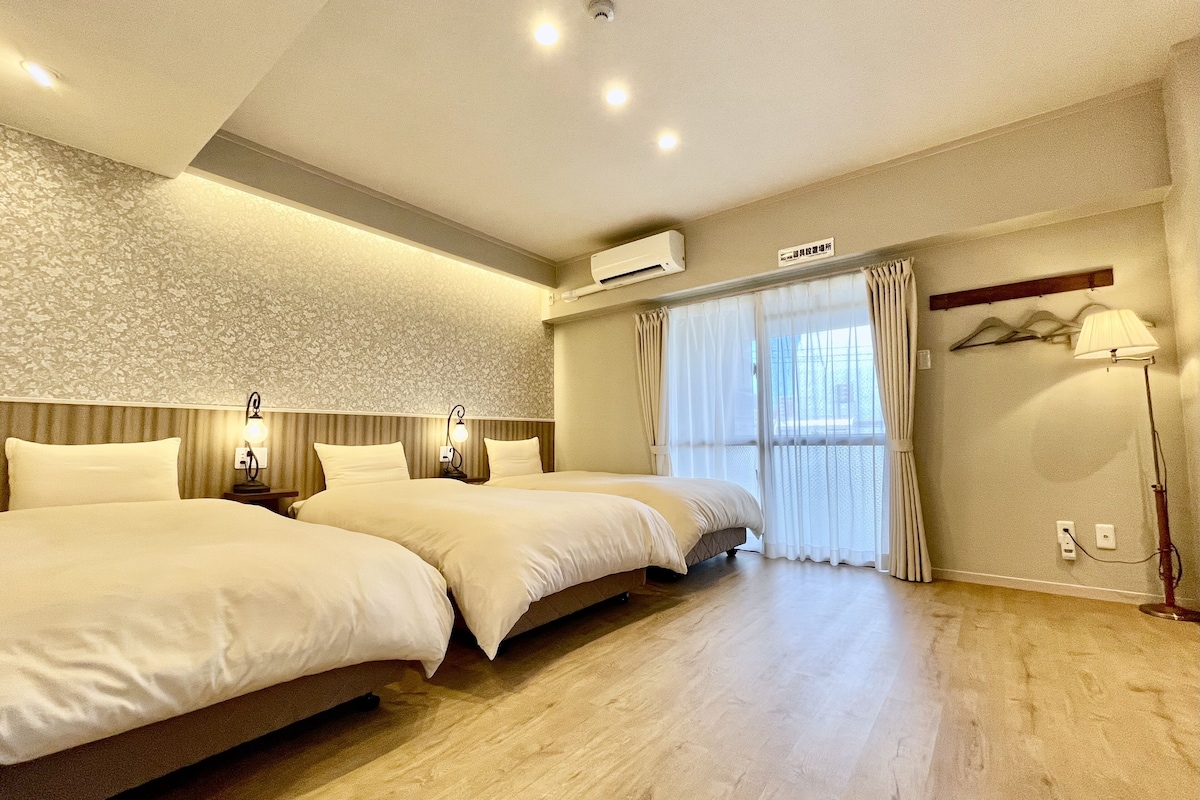 A hotel room with two beds, a large window with curtains, a ceiling light, and a wall-mounted air conditioner.