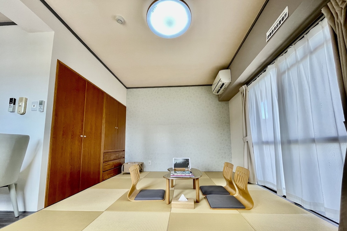 A room with tatami mats, a wooden wardrobe, a small table with a laptop, and a window with curtains.
