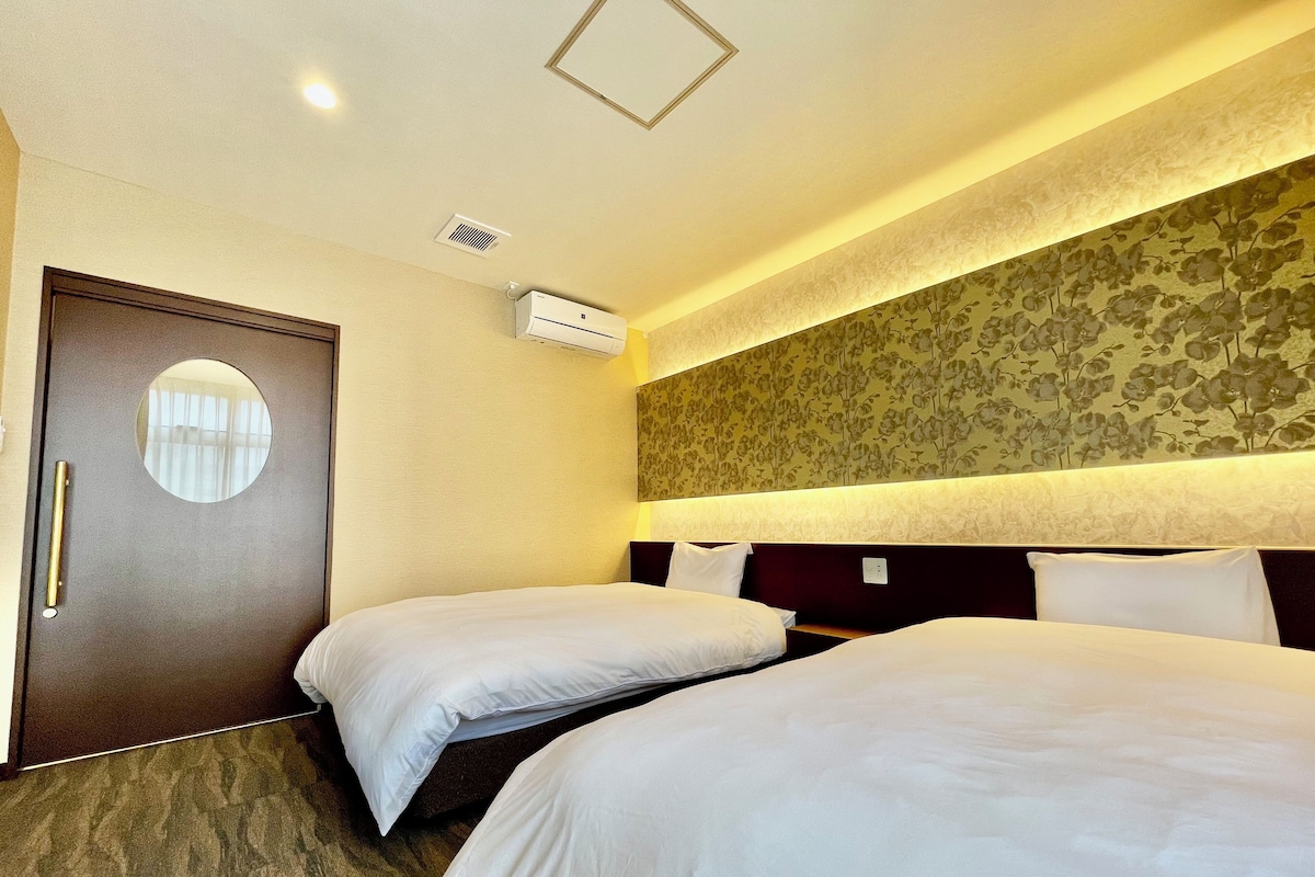 A hotel room with two beds, a round mirror, a door with a handle, and an air conditioning unit.