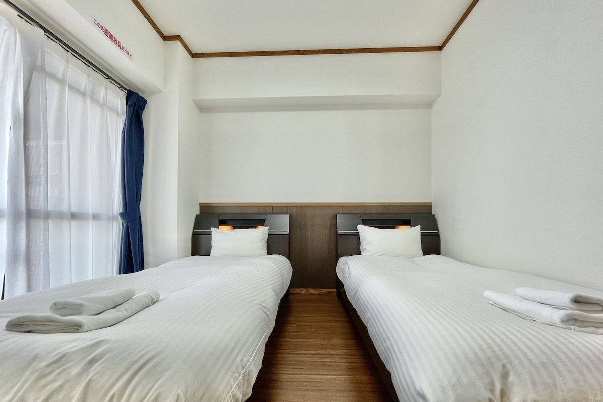 A hotel room with two beds, wooden flooring, white bedding, and a window with curtains.