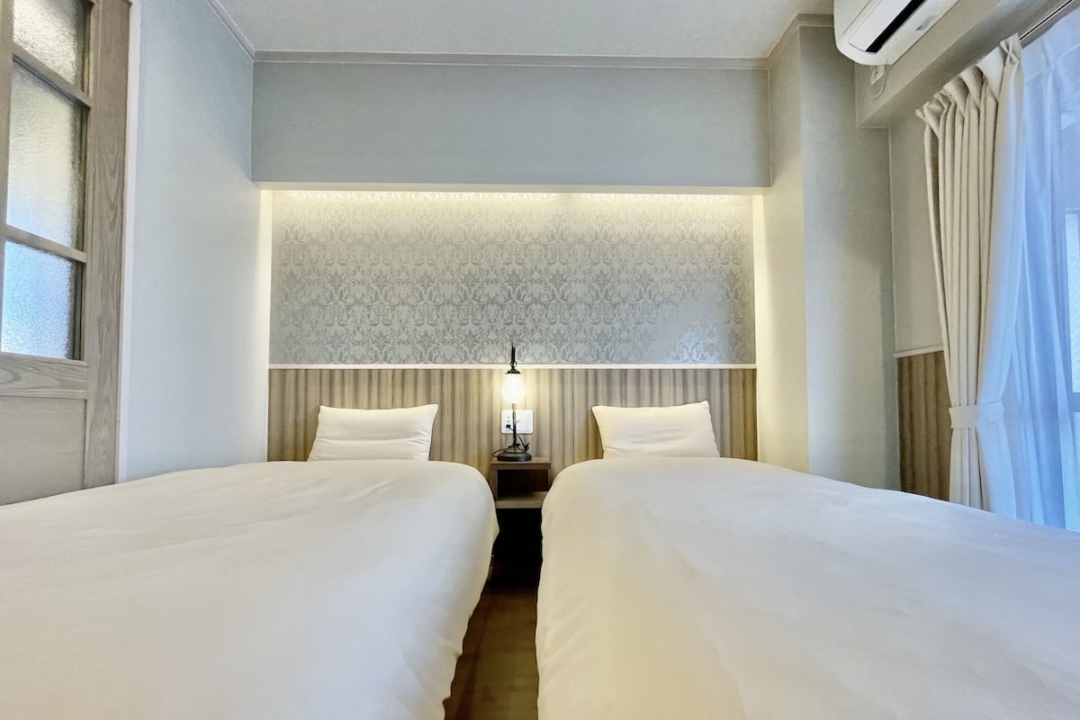 Ein Hotelzimmer mit zwei Betten, einem Nachttisch mit Lampe, einem gemusterten Wandteppich und einem Fenster mit Vorhängen.