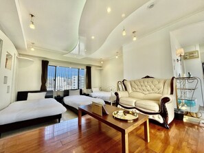 A modern hotel room with a large window, a sofa, two beds, a wooden coffee table, and a metal shelf. - Hotel Tenshi-Kan Kumoji (Naha)