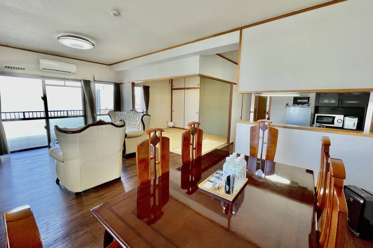 Ein modernes Hotelzimmer mit einer Couch, einem Esstisch mit Stühlen, einer kleinen Küchenzeile und einer Schiebetür zu einem Balkon.