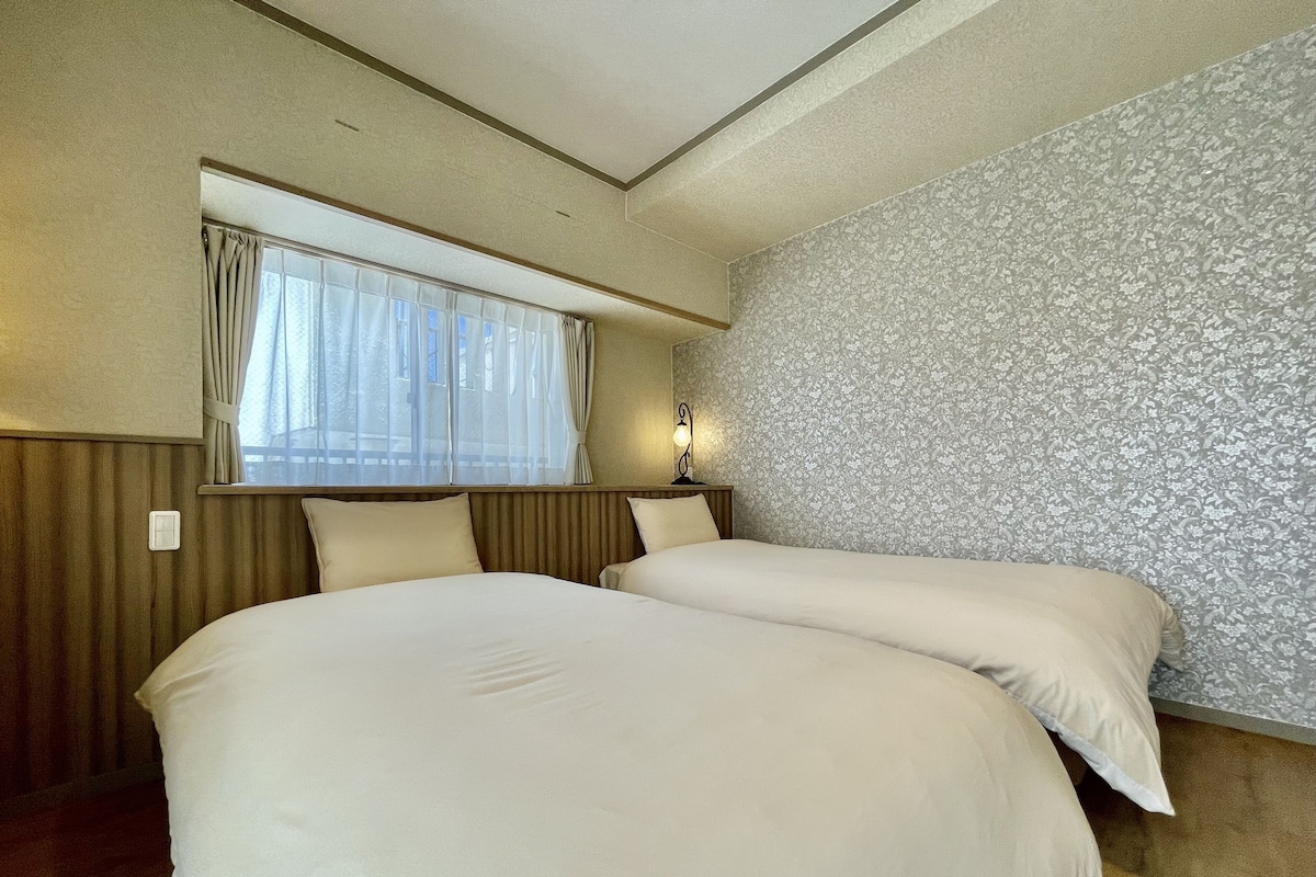 A hotel room with two beds, a window with curtains, a wall with a floral pattern, and a bedside lamp.