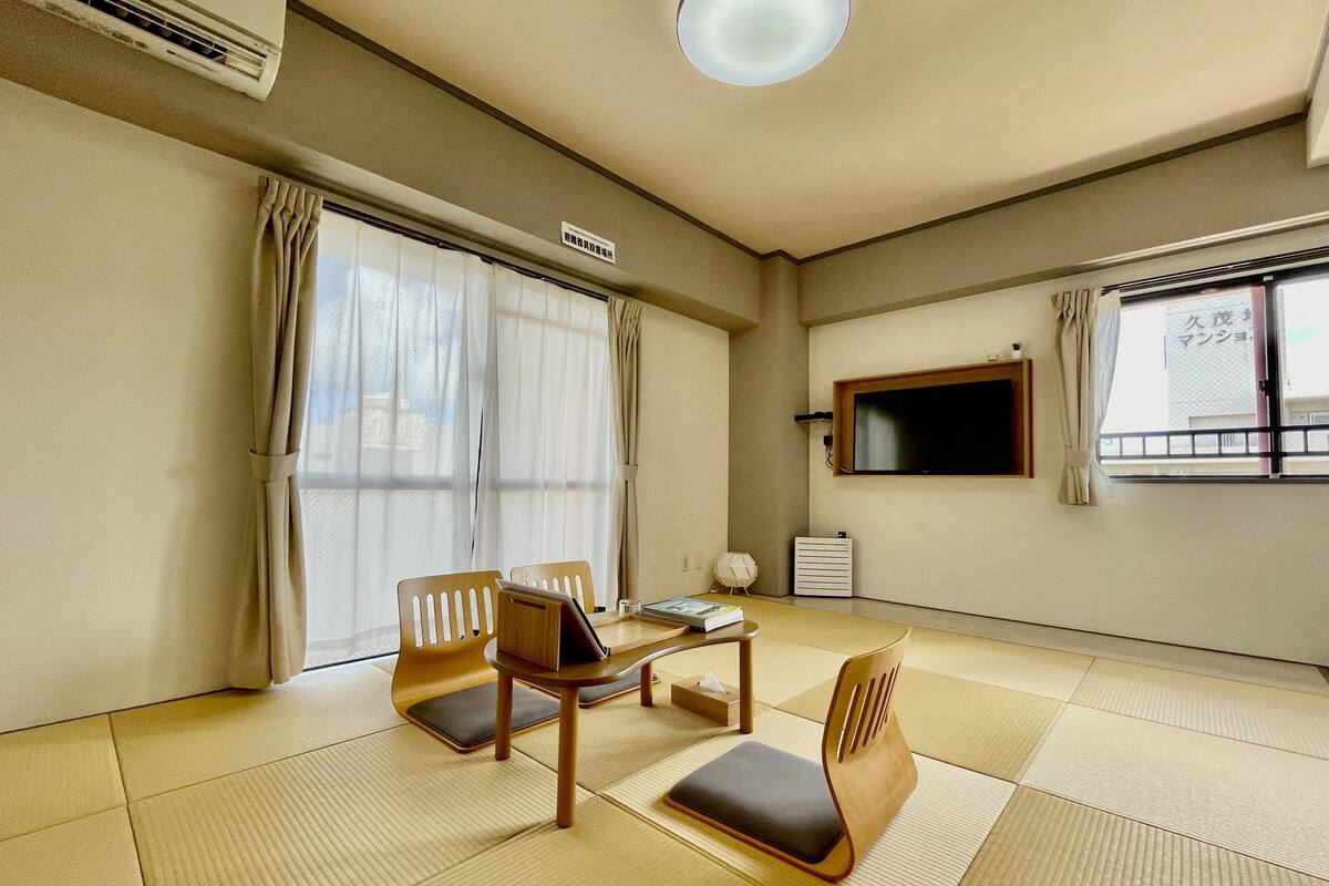Ein Zimmer mit Tatami-Böden, einem an der Wand befestigten Fernseher und einem fenster mit Vorhängen.