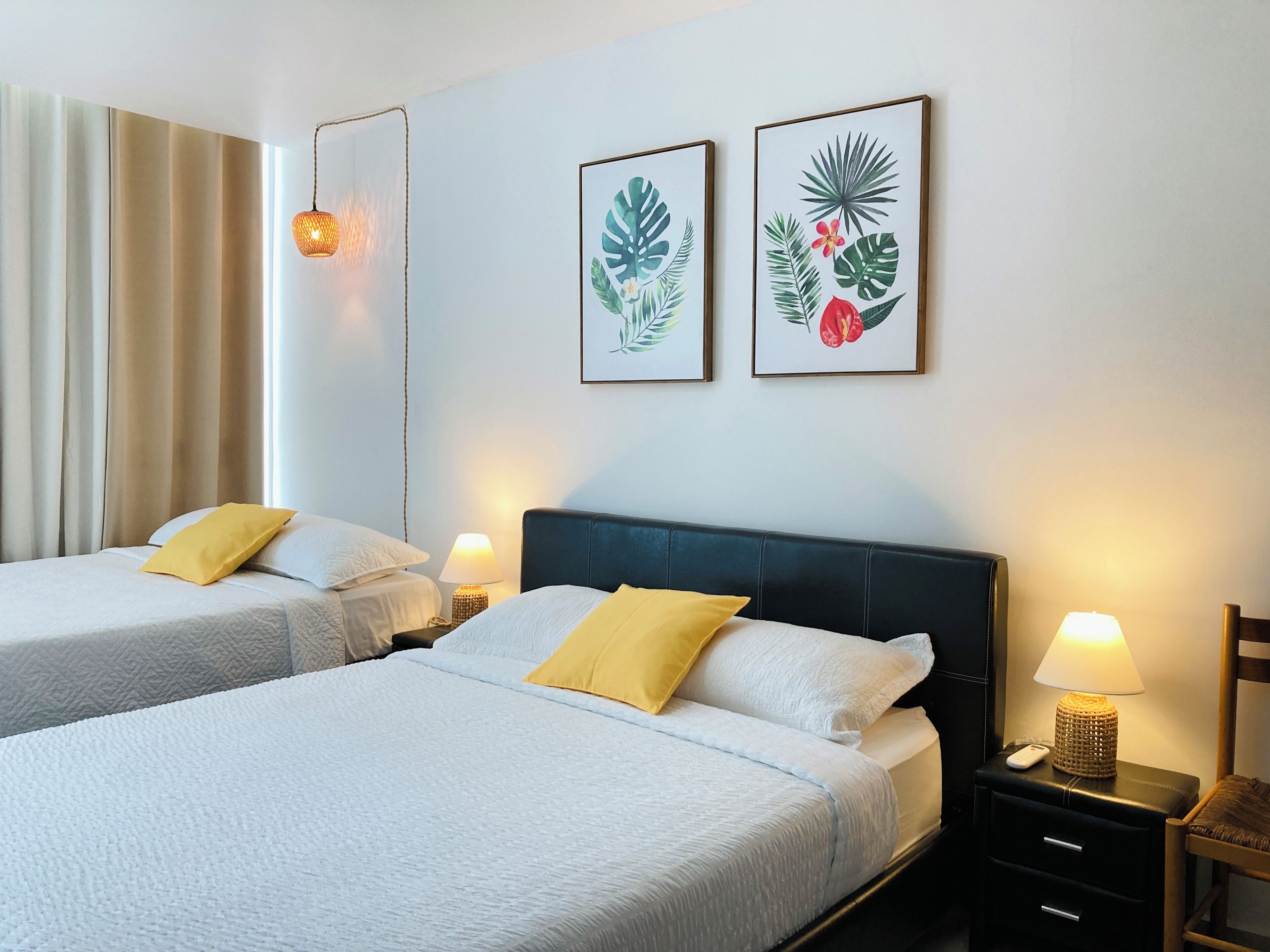 A hotel room with two beds, a nightstand with a lamp, and two framed botanical prints on the wall.