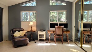 Una habitación con un sillón de cuero marrón, una mesita con una lámpara, una estufa antigua, un escritorio de madera con dos monitores y ventanas grandes con vistas a árboles.