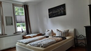 Een slaapkamer met een bed, een raam met gordijnen, een radiator, een inlijsting spiegel en een stadslandschap schilderij aan de muur.
